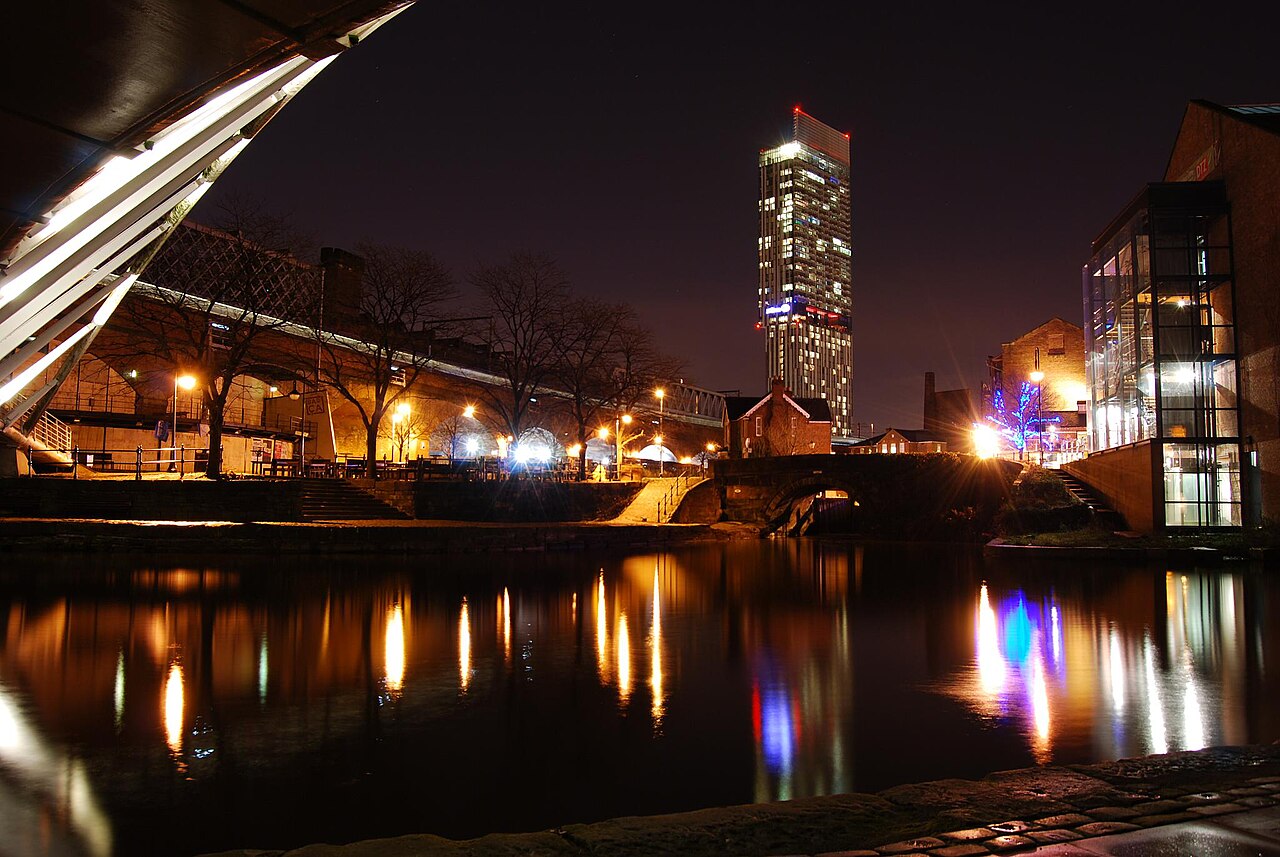 The canal basin at night, an angular footbridge lit up in the foreground, a stone arch bridge beyond it, and the lit tower of Beetham Tower on the skyline.