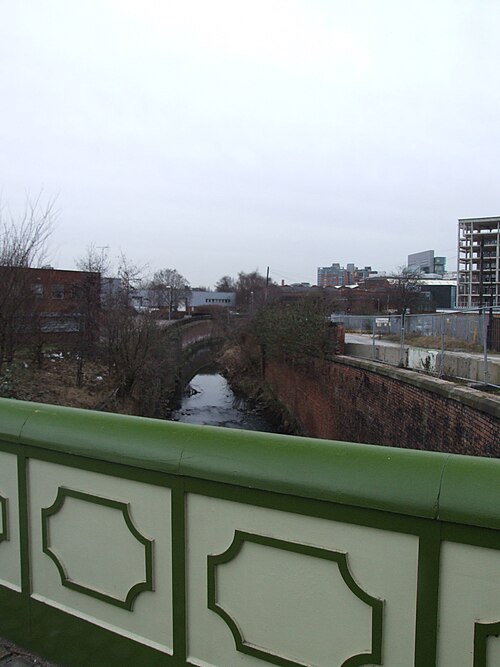 The River Medlock in a brick lined channel beneath a small stone arch bridge, seen from a green painted parapet with modern towers behind.
