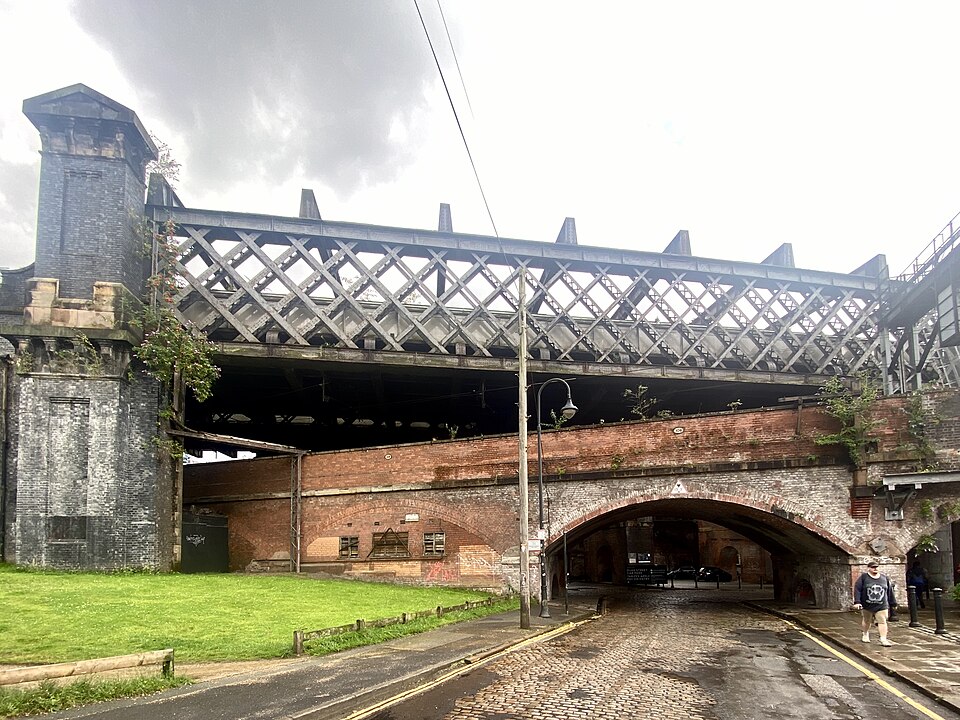 A cobbled street passes beneath two crossing viaducts, a steel lattice bridge above a brick arch span, with a small brick tower at the corner.