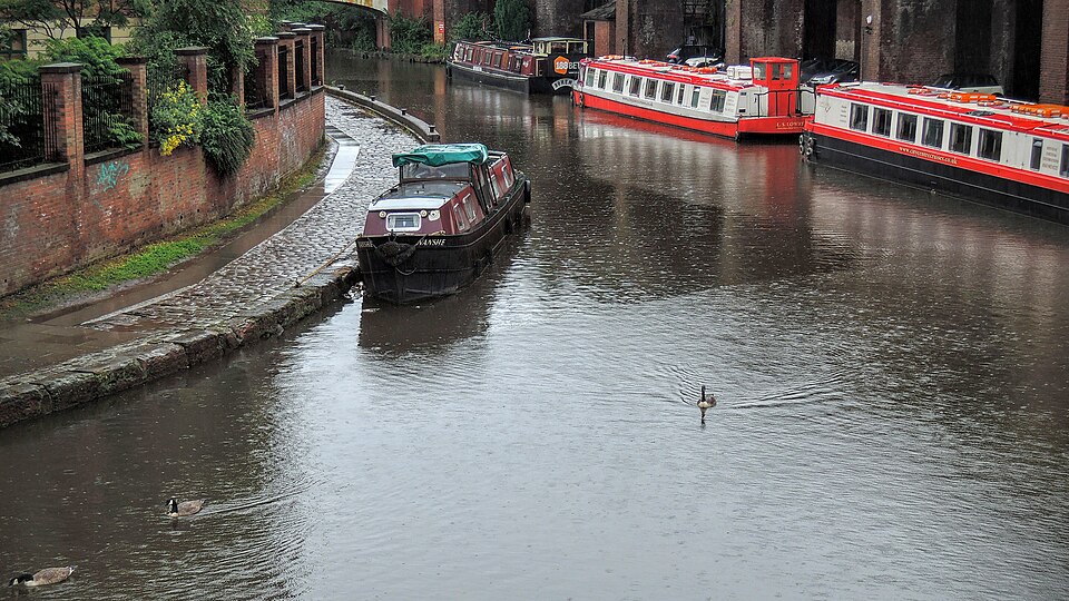 Narrowboats moored along a cobbled towpath in the canal basin, brick retaining walls on one side and a steel lattice footbridge crossing beneath the railway viaducts behind.