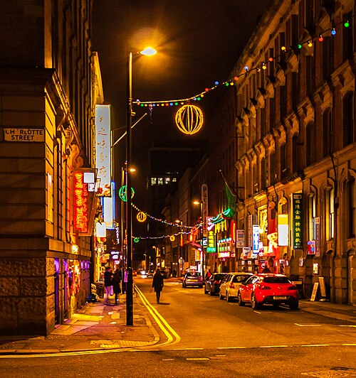 The Charlotte Street corner at night, strings of lit lanterns overhead and red and gold restaurant signs stacked up a stone fronted building.