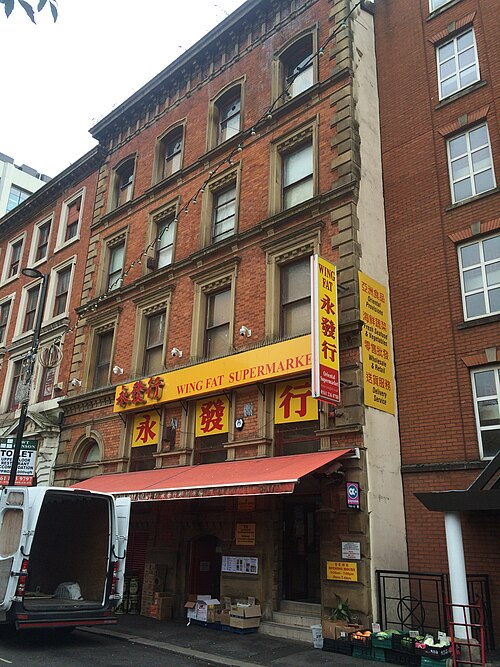 The Wing Fat Supermarket sign in red and gold running the width of a brown brick Victorian warehouse front, arched window heads stacked above.