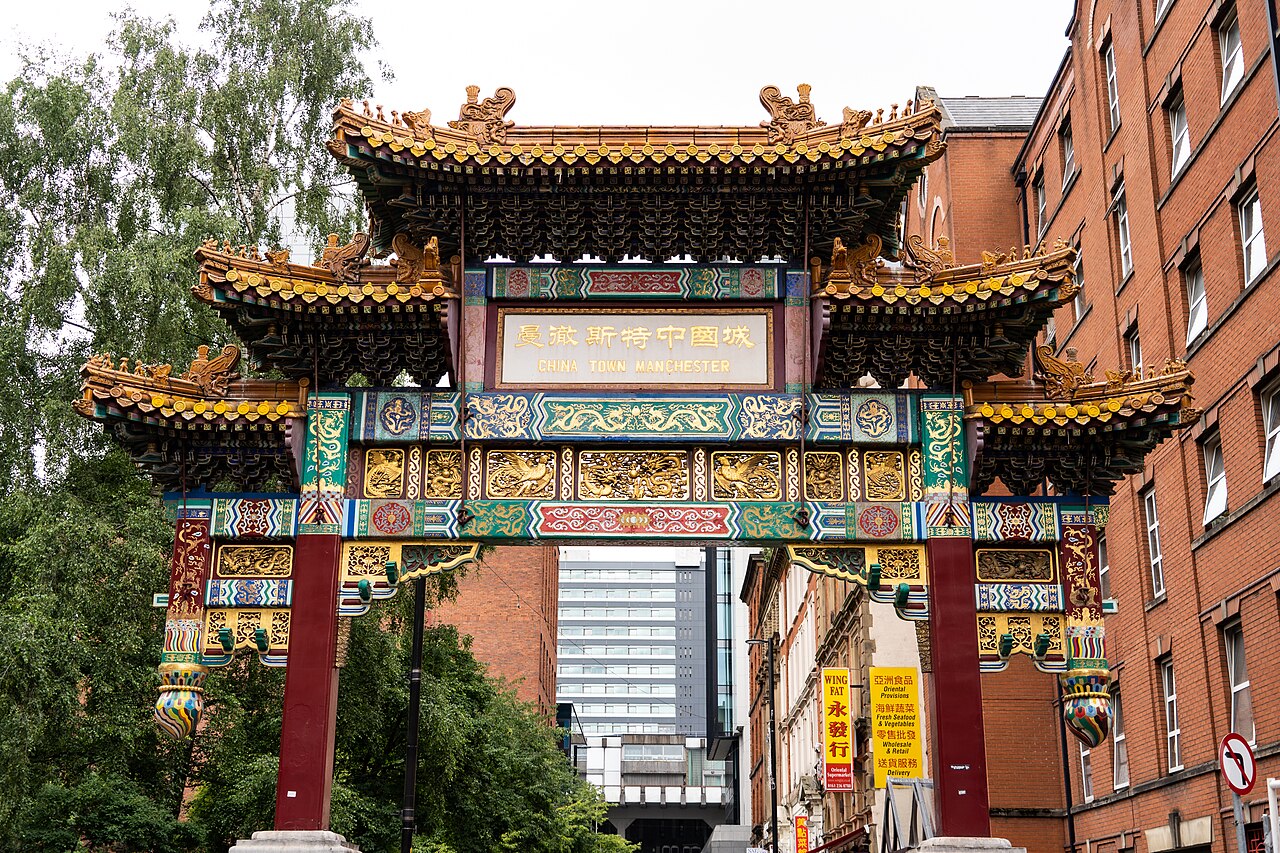 The painted archway close up, tiered gold roofs and red columns rising above a corner building carrying more restaurant signage.