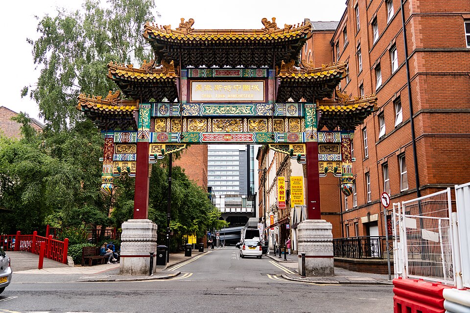 The arch and the Faulkner Street junction seen from the pavement, a red pedestrian barrier and trees to one side and a tower block visible through the opening. The streetscape immediately around the China Buffet Restaurant, which the Street View beside it shows directly.