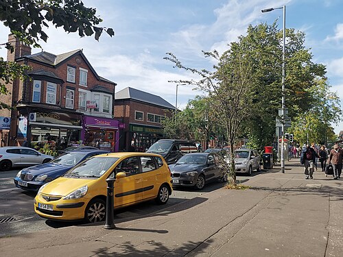 A row of red brick two storey buildings on Wilbraham Road, independent shopfronts at street level under bay windows and a gabled roof, cars parked along the kerb and pedestrians on the far pavement.