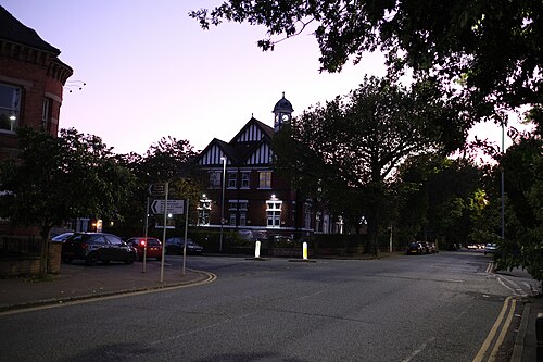 A black and white timber framed building with a corner clock tower, its windows lit at dusk, cars parked along the road and direction signs at the junction in front of it.