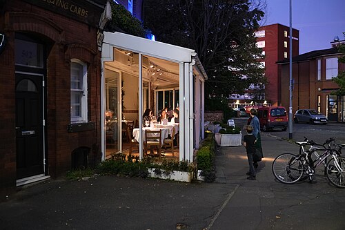 A red brick corner building with a lit glass fronted extension at night, a table laid inside, bicycles chained by the door and a tall apartment block behind.