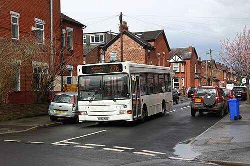 A single decker bus on the 276 route to Withington Hospital passes red brick terraced houses with bay windows and gabled dormers, cars parked along both kerbs and blossom on a street tree.