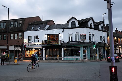 The corner at Barlow Moor Road, a cream painted Victorian building with dormer windows over a deli and an estate agent, a cyclist crossing at the lights.