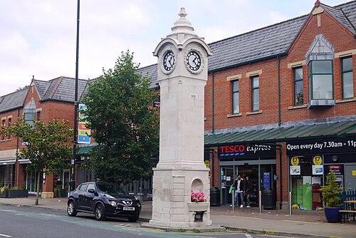 A Portland stone Victorian clock tower on the pavement outside a corner supermarket, red brick shopfronts with green gabled dormers behind.