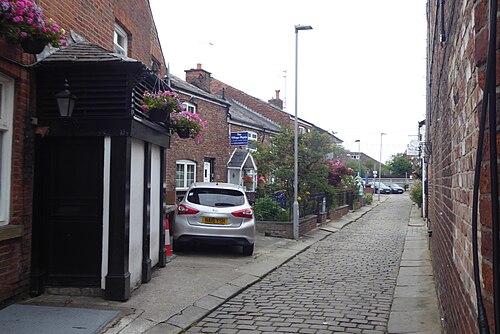 A cobbled lane behind the Wilmslow Road shops, red brick walls on both sides and hanging baskets over one doorway, a car parked along the kerb.