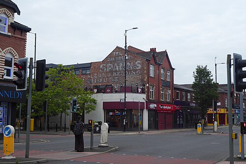 A faded painted sign for a bedding merchant high on a red brick gable at the Wilmslow Road junction, traffic lights and shopfronts below it.