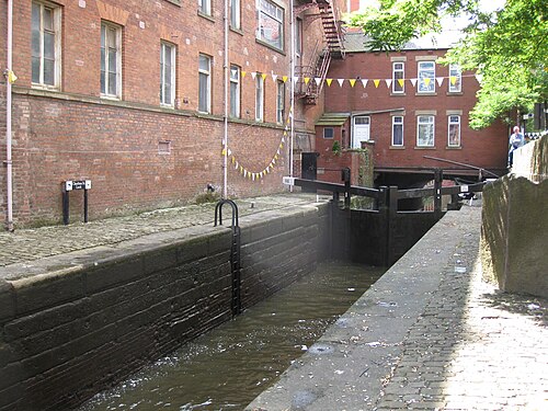 Chorlton Street Lock on the Rochdale Canal, a stone lock chamber with black timber gates, a cast iron ladder set into the wall and bunting strung across from the brick warehouse above.