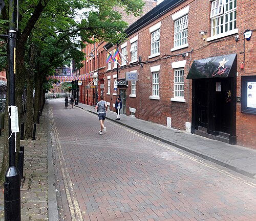 A row of red brick buildings with white sash windows, a black shopfront with neon star signage on the right and a hotel sign over an arched doorway further along, brick paving and street trees in front.