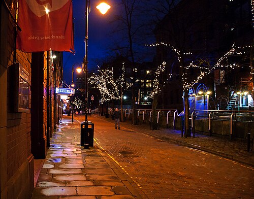 The canalside walk at night, string lights woven through bare trees, wet cobbles reflecting orange lamp light and a Manchester Pride flag hanging from a building on the corner.