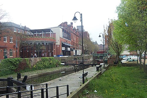 A canal basin with a curved colonnaded building on the left and taller red brick buildings behind it, a stone towpath and black iron railing beside the water, and a footbridge crossing the canal further along.