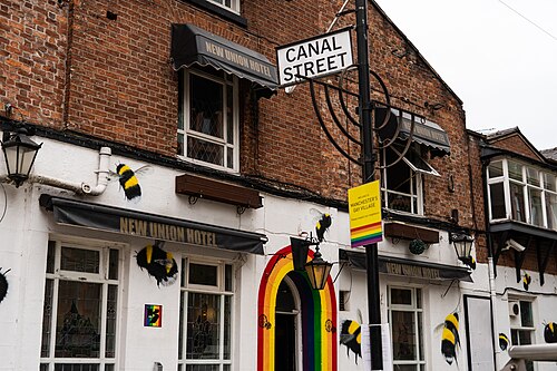 A Canal Street sign mounted on a lamp post outside the New Union, a red brick building with a whitewashed ground floor, a rainbow arch over its doorway and bee decorations fixed to the wall.