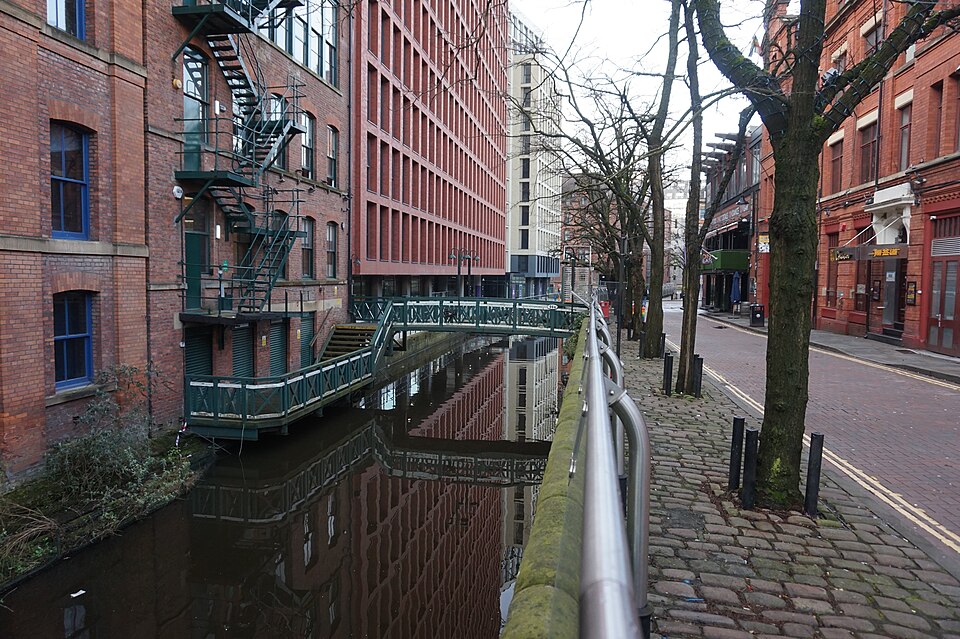 A red brick canalside building with cast iron fire escapes climbing its wall, a green painted footbridge crossing the canal in front of it, and cobbles either side of the towpath.