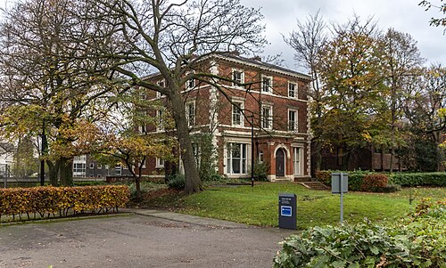 A three storey red brick Victorian villa with stone window surrounds and a pedimented doorway, set behind a lawn and mature trees, now a University of Manchester hall of residence.