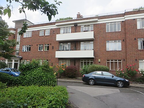 An Art Deco block of flats on Wilmslow Road in red brick with white rendered balconies and steel framed windows, rose bushes along the front path.