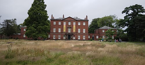 Platt Hall, a red brick Georgian mansion with a pedimented doorway and tall sash windows, standing behind rough meadow grass in Platt Fields Park.