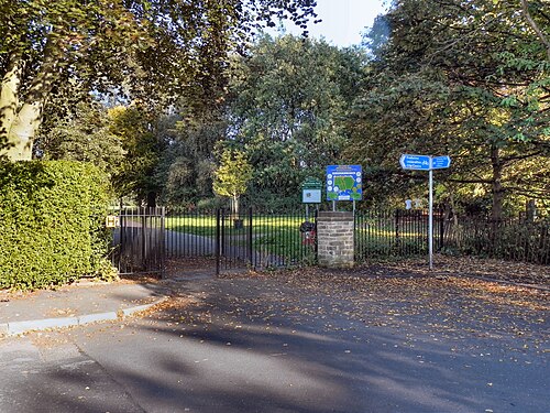 The iron gated entrance to Birchfields Park, a hedge and mature trees either side of the path and a park map fixed to a signboard.