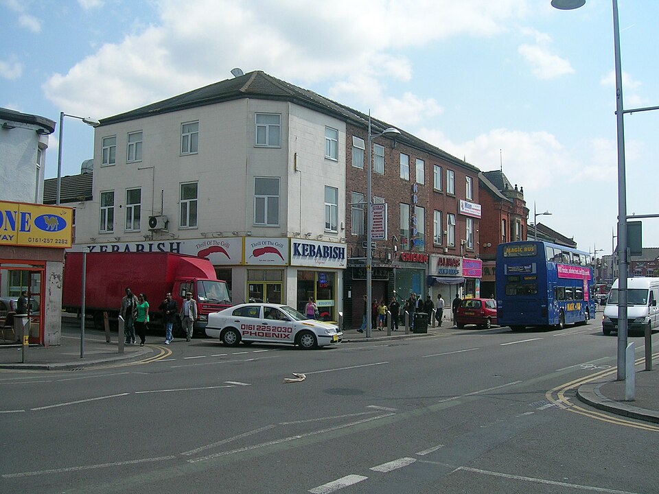 A corner block on Wilmslow Road with Kebabish and Alina's Jewellers on the ground floor, a Magic Bus double decker passing and pedestrians outside the shops.