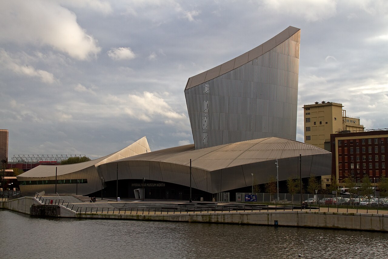 Imperial War Museum North's aluminium clad shards angled against the sky, dock water in the foreground.