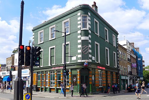 A three storey corner pub on Chalk Farm Road painted dark green, with a tiled ground floor and shopfronts running away down both streets.
