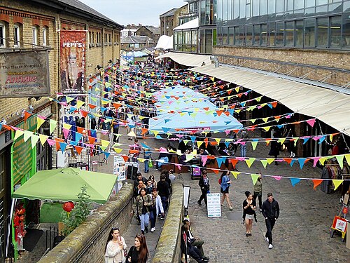 The Stables Market from above, a cobbled lane between brick buildings with stall canopies down both sides and bunting strung across it.