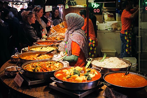 A food stall inside the market, eight steel pans of curry along the counter and a server working behind them.
