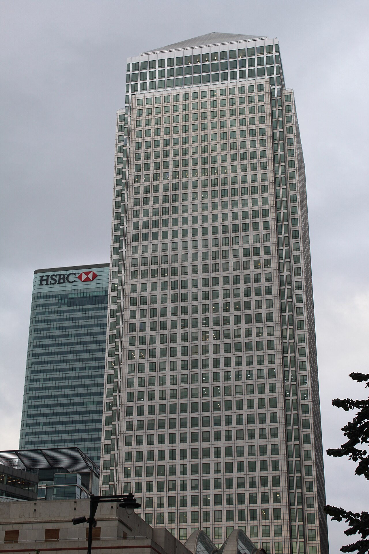 One Canada Square from the foot of the tower, its pyramid top against a grey sky, the HSBC building beside it and curved glass canopies at street level.