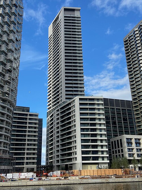 New flats at Wood Wharf across the water, a tall tower with continuous balconies behind lower blocks, builders' hoarding along the quay.