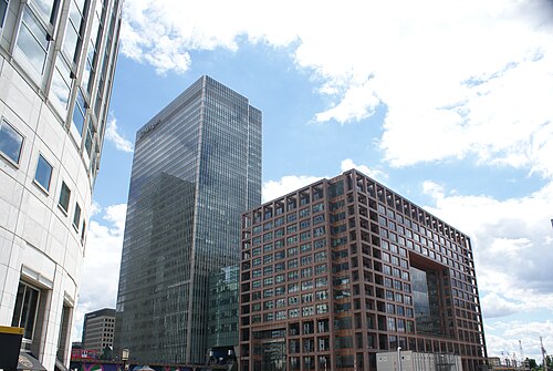 Seen from Mackenzie Walk, a dark glass office tower beside a brown steel framed building with a rectangular opening cut through its upper floors, and a DLR train passing at the left edge.