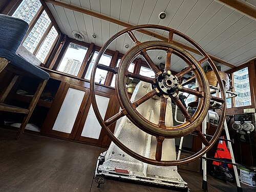 The wheelhouse of a moored ship, a wooden ship's wheel on a white pedestal, varnished panelling and Canary Wharf towers through the windows.