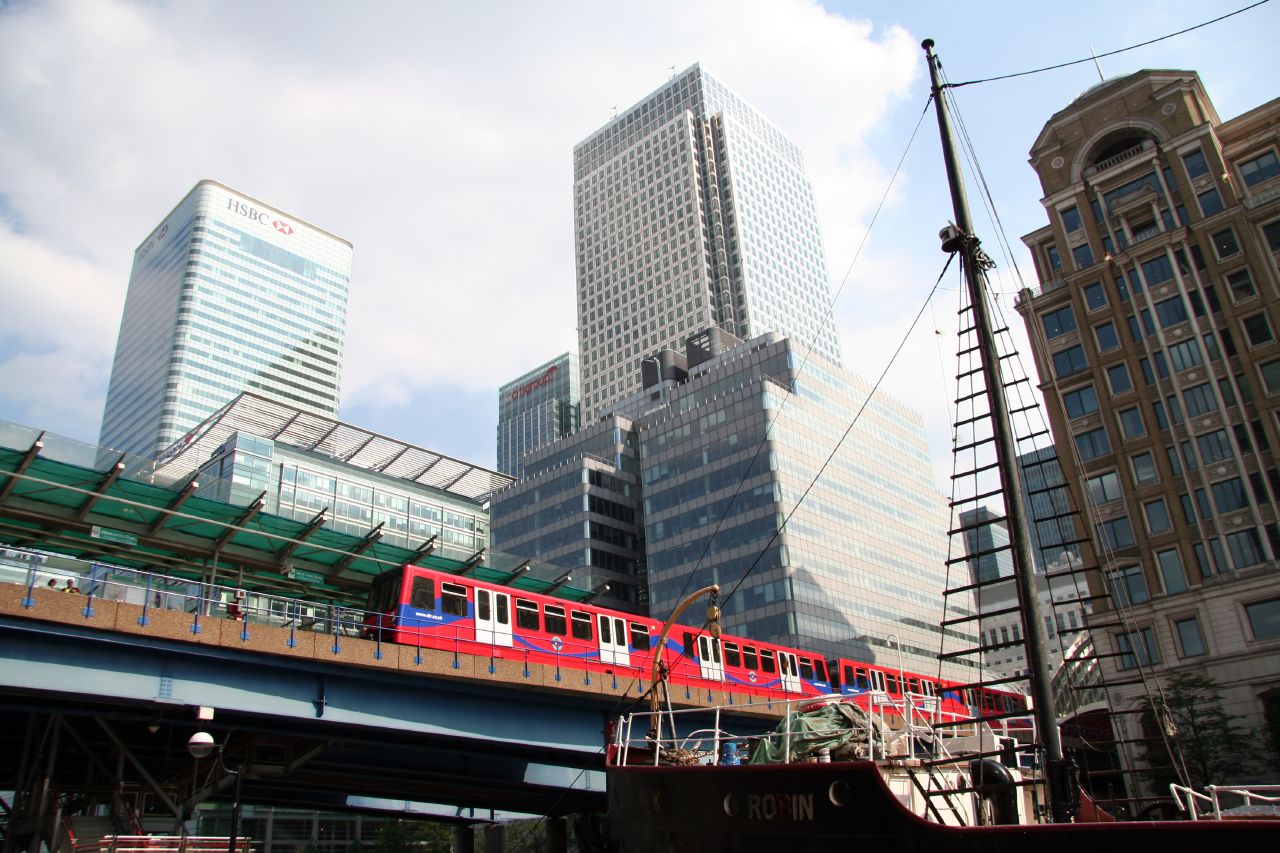 A red DLR train on the viaduct over the dock, the Canary Wharf office towers behind it and the masts of the moored steamship SS Robin in the foreground.