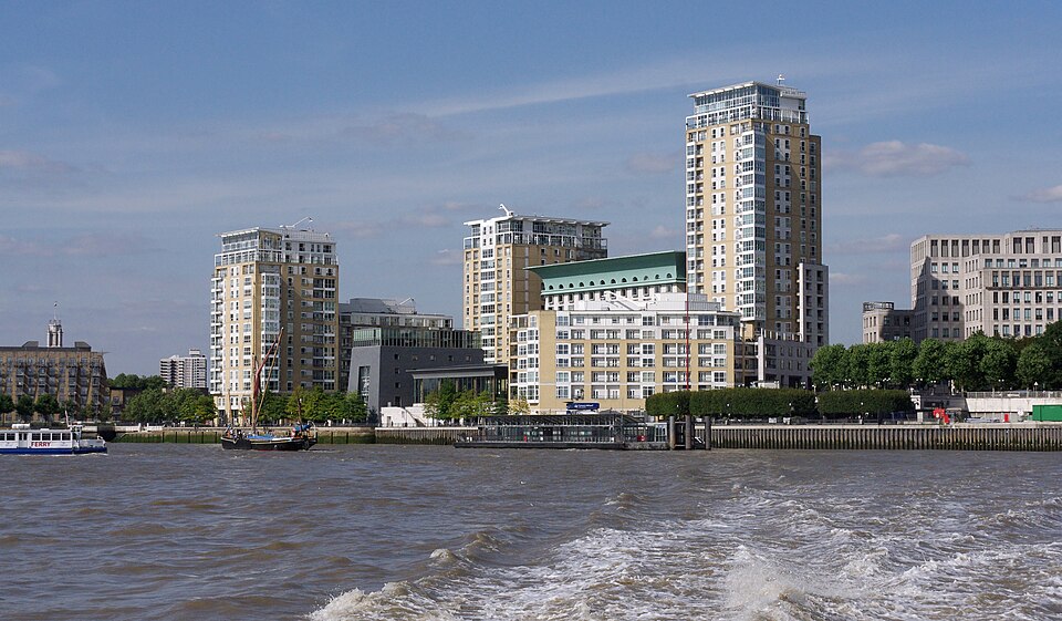 The Thames frontage at the west end of Canary Wharf, apartment blocks along the water with a lower building under a green roof, a sailing barge and a pier in front of them.