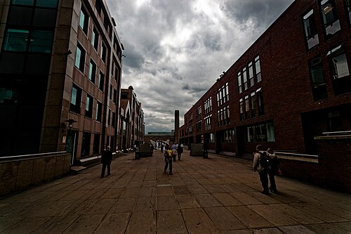 Peter's Hill looking south towards the river, a wide paved route between granite and brick offices, with the chimney of Tate Modern showing at the end of it under a dark sky.
