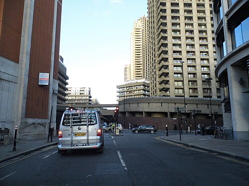 Long Lane at the Barbican, a van and cars on the road, one of the Barbican towers on the right and a pedestrian bridge crossing the street ahead.