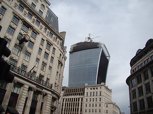 The Walkie Talkie seen from Cannon Street while it was still topped by a crane, above a stone office building with a projecting clock over the pavement.