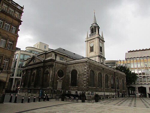 St Lawrence Jewry seen across Guildhall Yard, the clock tower and lead spire above a stone flank wall, with the patterned paving of the yard in front.