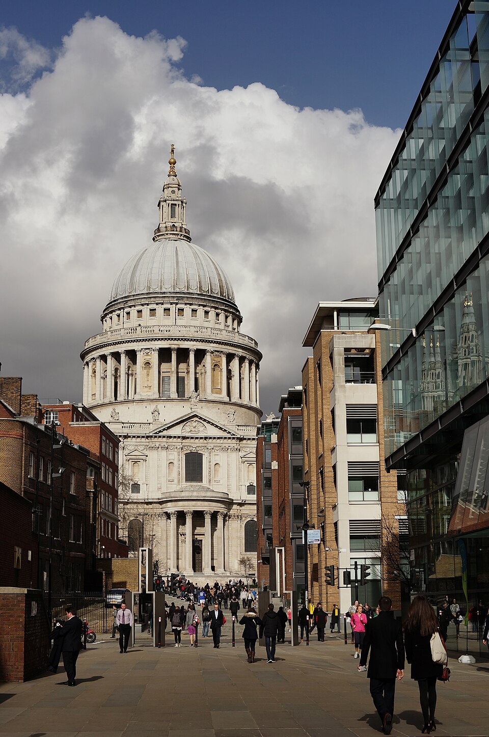 St Paul's Cathedral seen up Peter's Hill from the south, the dome above the transept portico, brick and glass offices along both sides of the paved route and people walking up it.