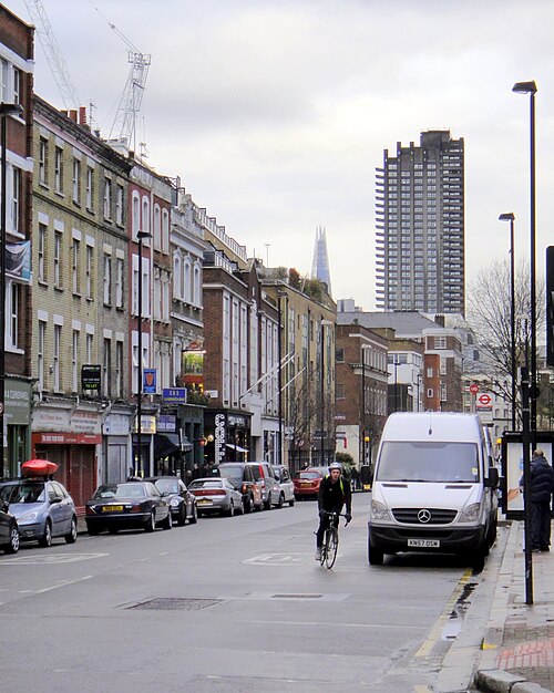 Goswell Road looking south, shopfronts and stuccoed upper floors along the left side, with a Barbican tower and the top of the Shard in the distance.