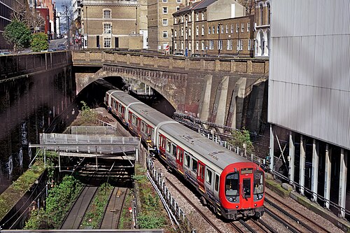An Underground train coming out of the tunnel into the open railway cutting near Farringdon, brick retaining walls on both sides and buildings standing above them.