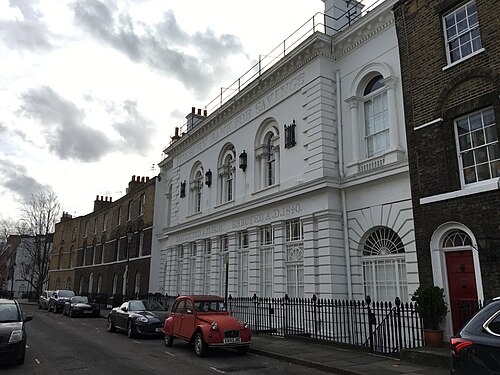 The former Finsbury Bank for Savings on Sekforde Street, a white stuccoed front lettered INSTITUTED A.D.1816 ERECTED A.D.1840, with a brick terrace running away behind it and an orange 2CV parked outside.