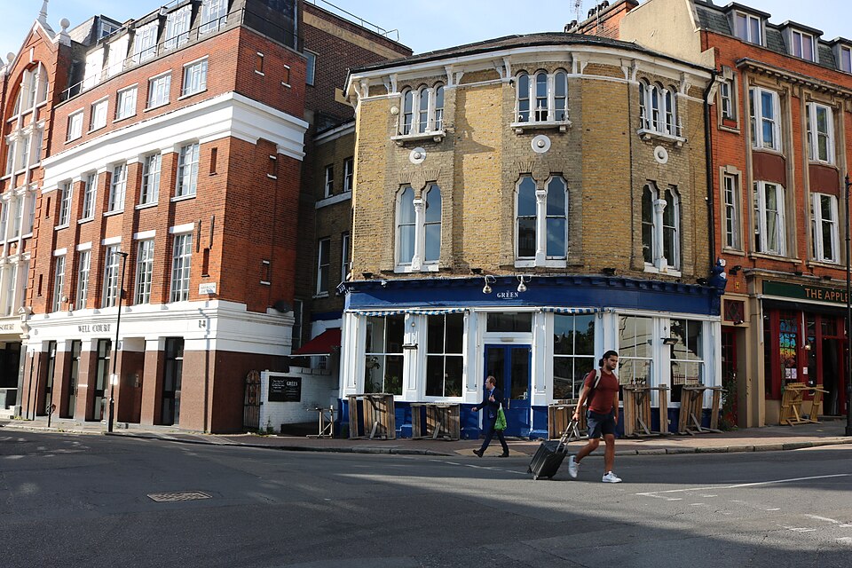 The west corner of Clerkenwell Green at Farringdon Lane, a yellow brick building with a blue painted shopfront on the ground floor and the red brick block called Well Court beside it.