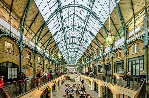 The Central Avenue inside the market building, green painted iron arches under a glazed roof, yellow brick walls with arched shopfronts, hanging lanterns and cafe tables on the sunken floor below.