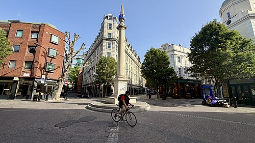 The junction at Seven Dials early on a summer morning, the stone column on its stepped base in the middle of a setted circle with streets running off it on all sides.