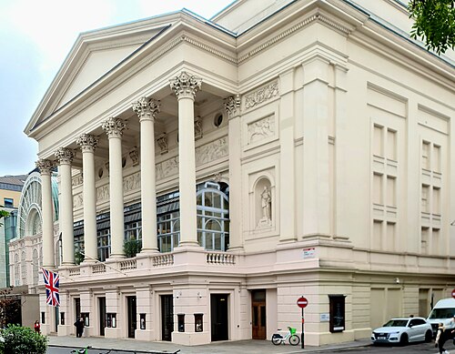 The Bow Street front of the Royal Opera House, six Corinthian columns under a pediment with a carved frieze behind them, and the iron and glass of the Floral Hall along the left.