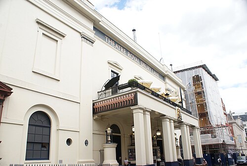 The Theatre Royal Drury Lane seen along Catherine Street, a cream stucco front with the name across the parapet and a colonnaded entrance under a first floor terrace.